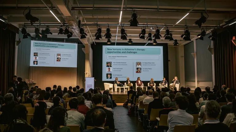 View from the back of a conference room towards the stage; the stage is lit, the people in the audience are sitting in the dark.