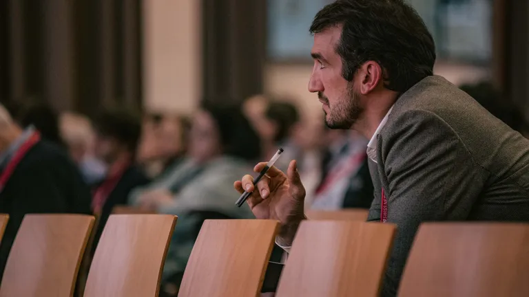 A man sits in the audience, looking intently ahead, playing with a pen in his right hand; the row of seats in front of him is empty.