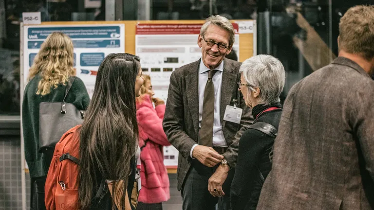 A man and two women are standing in a room with a poster exhibition, chatting and laughing. They are engrossed in conversation.