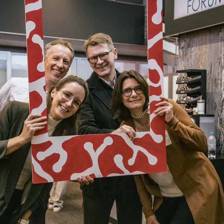 Four persons are standing behind a large Polaroid picture frame, holding it in their hands and smiling at the camera.
