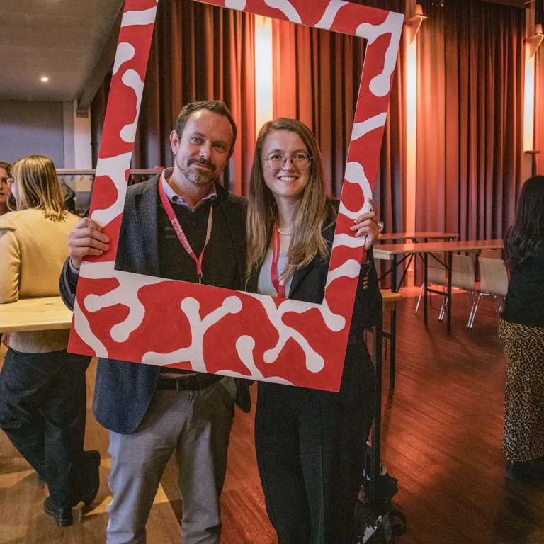 A man and a woman pose with a life-size red and white Polaroid picture frame and smile at the camera.