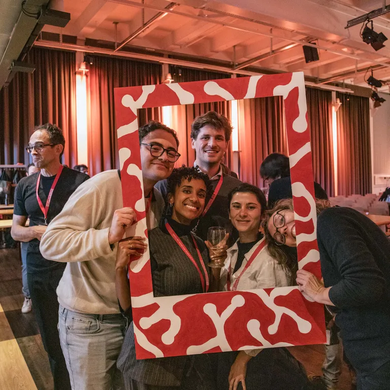 A group of five people pose behind a large red and white Polaroid frame and smile at the camera.