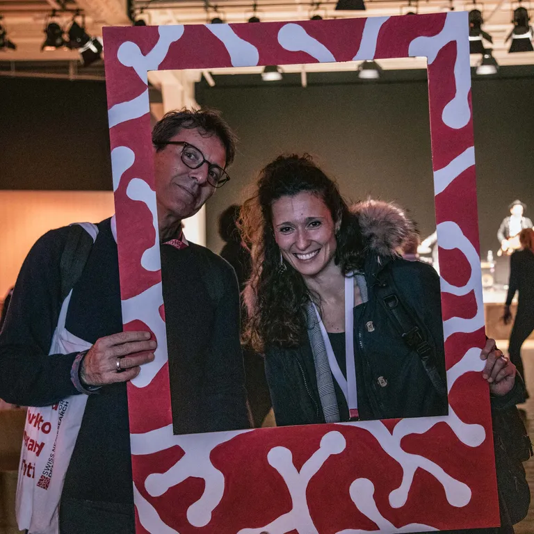 Two people are standing behind a large Polaroid picture frame, holding it in their hands and smiling at the camera.