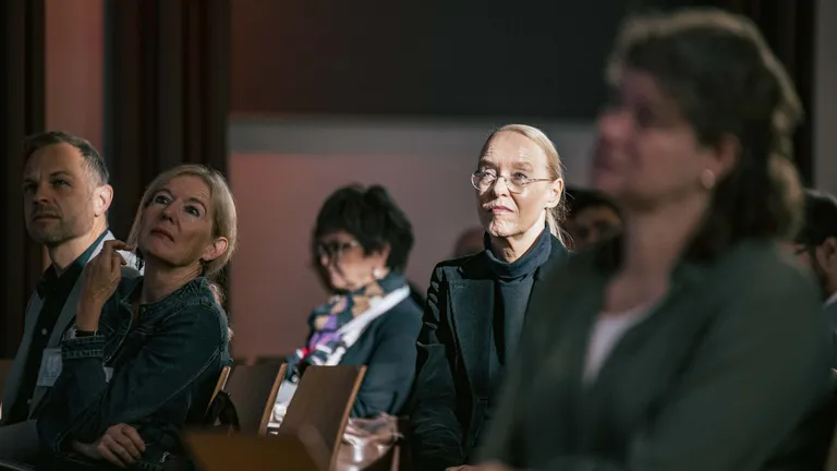 Perspective of 5 individuals listening to a conference. The focus is on a woman in the centre of the image, who is being illuminated by the sun.