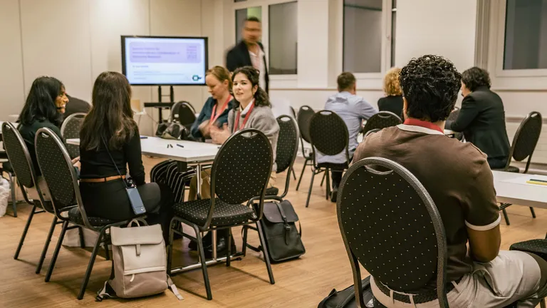 A room with tables and chairs arranged in a workshop setup. People are sitting on some of the chairs.